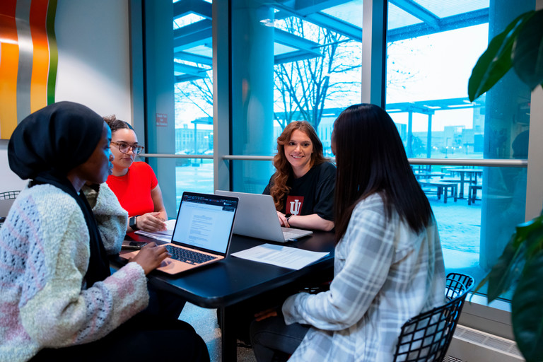 Students sit at a table with their laptops open and talk.