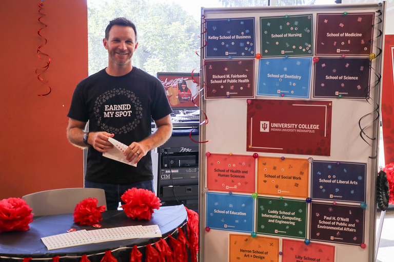 A man stands behind a table wearing a shirt that says "Earned My Spot". He is standing next to a board that has flyers for each of the colleges in University College.